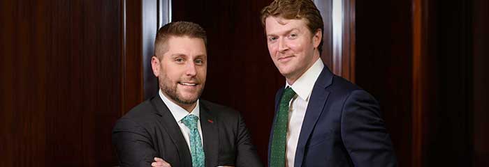 Attorneys Aaron Burke and Rob Bogdanowicz standing in business suits in front of a set of wooden doors.