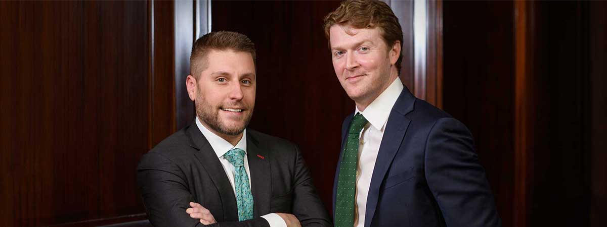 Attorneys Aaron Burke and Rob Bogdanowicz standing in business suits in front of a set of wooden doors.