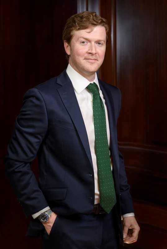 Attorney Aaron Burke in a blue suit with a green tie standing next to a wooden door.